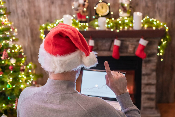 Man freelancer in santa claus hat working on laptop with graphs and charts raising a finger up sitting near christmas tree and fireplace. - Valokuva, kuva