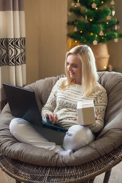 Registro de entrega en línea. Mujer se sienta en silla redonda, sosteniendo una caja de regalo en su mano y hace la entrega de regalos de Navidad en línea utilizando el ordenador portátil. - Foto, imagen
