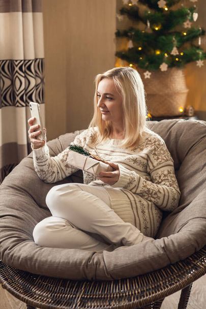 Online registrace zásilky. Žena sedí v kulaté židli, drží dárkovou krabici v ruce a dělá dodání vánočních dárků on-line pomocí smartphonu. - Fotografie, Obrázek