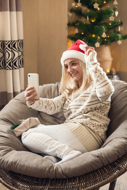 Saludos en línea de Navidad y Año Nuevo. Mujer se sienta en silla redonda en Santa sombrero y saludos seres queridos en línea utilizando el teléfono inteligente. - Foto, imagen