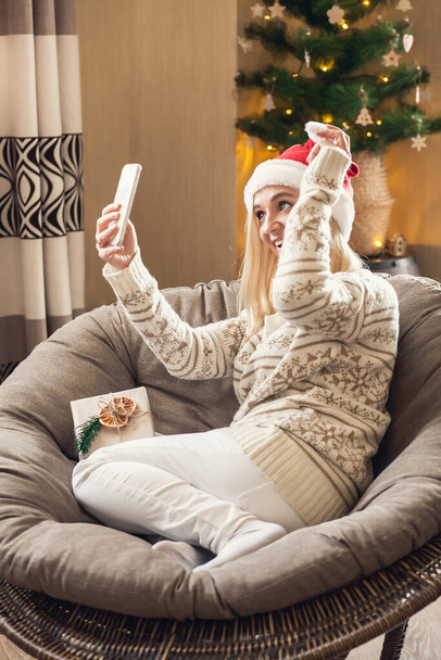 Online vánoční a novoroční pozdravy. Žena sedí v kulaté židli v klobouku Santa a pozdravy milované on-line pomocí smartphonu. - Fotografie, Obrázek