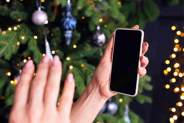 egy nő kezet lóbál az okostelefon képernyőjén egy karácsonyfa hátterével szemben. Az online üdvözlés fogalma. koronavírus és karantén az új évre. üres hely a tervezéshez - Fotó, kép