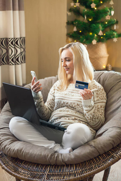 Online vánoční a novoroční nakupování - žena sedí na kulaté židli a platí online nákupy kreditní kartou pomocí notebooku a smartphonu. - Fotografie, Obrázek