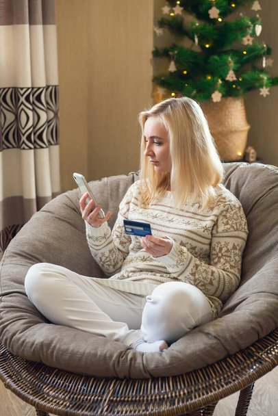 Online vánoční a novoroční nakupování - žena sedí na kulaté židli a platí online nákupy kreditní kartou pomocí notebooku a smartphonu. - Fotografie, Obrázek