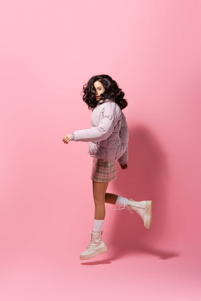 side view of brunette young woman in stylish winter outfit jumping on pink background - Foto, Imagen