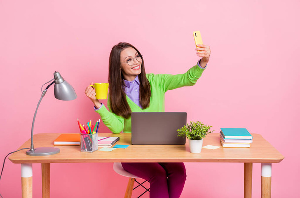 Photo of positive high school blogger girl sit table hold mug beverage make selfie smartphone isolated pink color background - Photo, Image