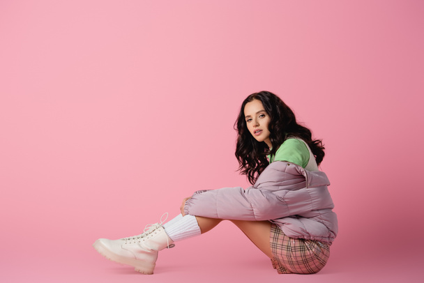 side view of brunette young woman in stylish winter outfit posing on floor on pink background - Foto, immagini