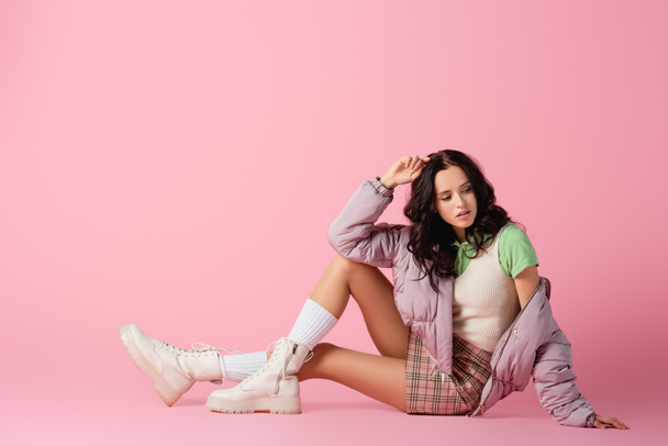 side view of brunette young woman in stylish winter outfit posing on floor on pink background - 写真・画像