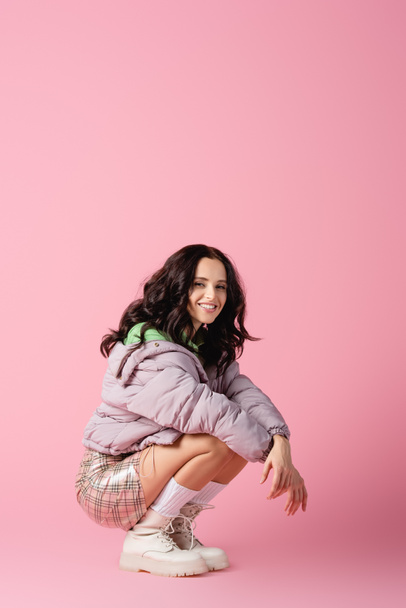 happy brunette young woman in stylish winter outfit posing on floor on pink background - Φωτογραφία, εικόνα
