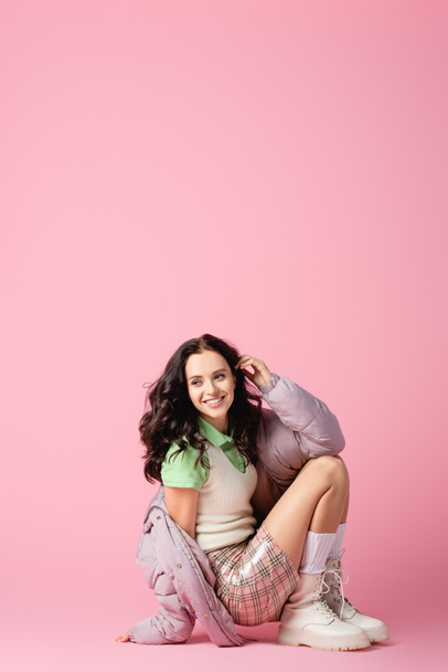 happy brunette young woman in stylish winter outfit posing on floor on pink background - Foto, immagini