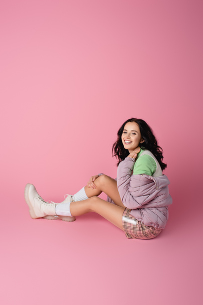 smiling brunette young woman in winter outfit posing on floor on pink background - Foto, afbeelding