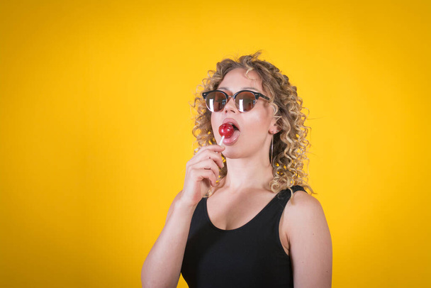 Beautiful amazing sexy smiling woman in sunglasses with a lollip, yellow background, studio shot - Fotografie, Obrázek