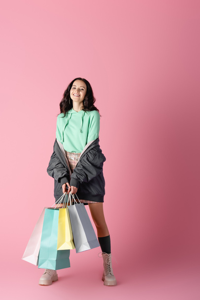улыбающаяся брюнетка молодая женщина в повседневном зимнем наряде с пакетами покупок на розовом фоне - Фото, изображение