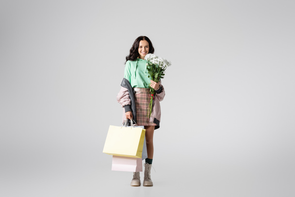 mosolygós barna fiatal nő elegáns téli ruha pózol virágok és bevásárló táskák szürke háttér - Fotó, kép