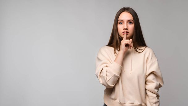 Young beautiful brunette woman wearing casual beige hoodie asking to be quiet with finger on lips. Silence and secret concept. Copy space for your text - Фото, изображение