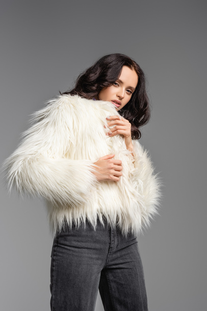 elegant brunette young woman in stylish white faux fur jacket posing on grey background - Zdjęcie, obraz