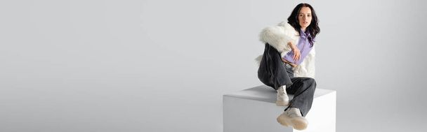 elegant brunette young woman in stylish faux fur jacket posing on cube on white background, banner - Foto, imagen