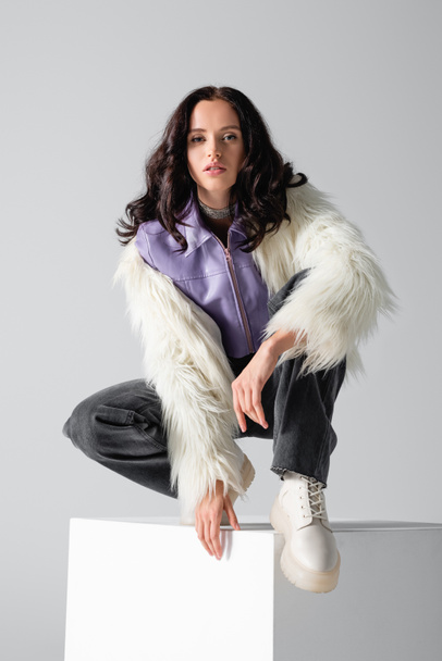 elegant brunette young woman in stylish faux fur jacket posing on cube on white background - Foto, Imagen