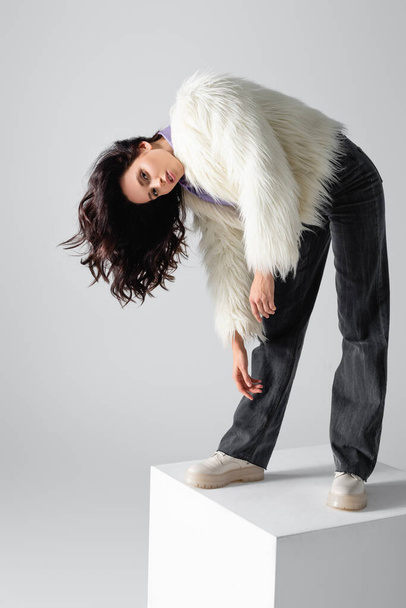 elegant brunette young woman in stylish faux fur jacket posing on cube on white background - 写真・画像