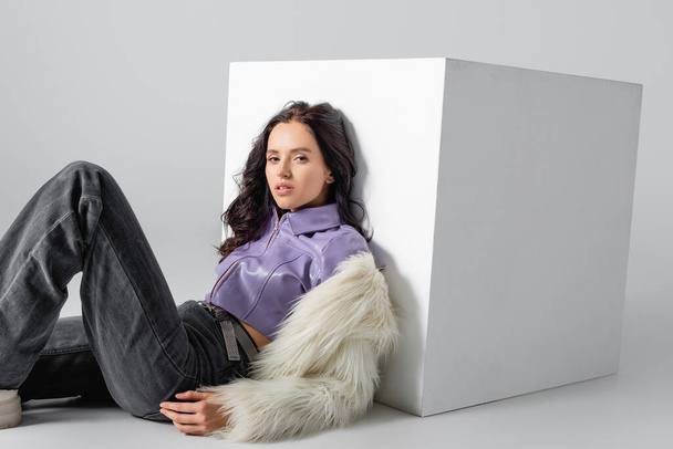 elegant brunette young woman in stylish faux fur jacket posing near cube on white background - Zdjęcie, obraz
