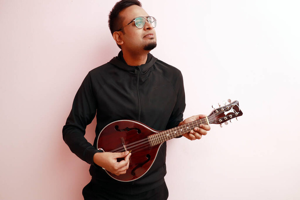 an stylish indian musician with goatee in black dress playing mandolin and looking up in light colored background - Foto, Imagen
