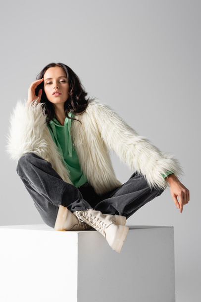 brunette young woman in faux fur jacket posing on cube on white background - Φωτογραφία, εικόνα