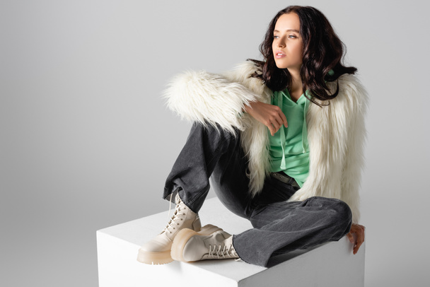 brunetka młoda kobieta w faux futro kurtka pozowanie na sześcian na białym tle - Zdjęcie, obraz