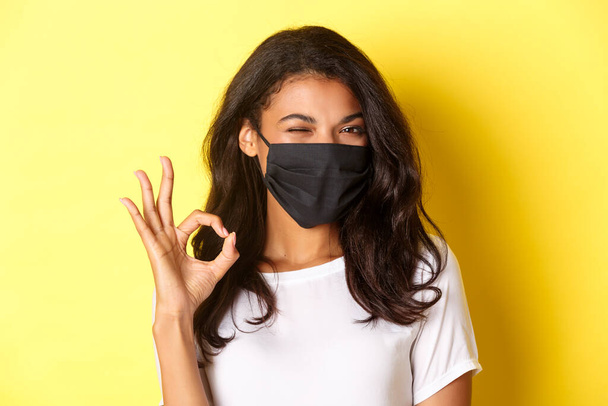 Koronaviruksen, pandemian ja elämäntavan käsite. Lähikuva röyhkeä afrikkalais-amerikkalainen tyttö musta kasvot naamio, osoittaa kunnossa merkki ja silmää, suositella jotain, keltainen tausta - Valokuva, kuva