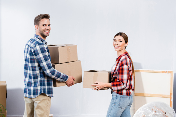 happy young couple looking at camera while holding cardboard boxes at home - Photo, Image
