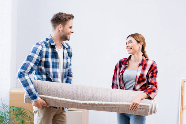 feliz pareja joven mirándose mientras sostiene el rollo de alfombra en casa - Foto, Imagen