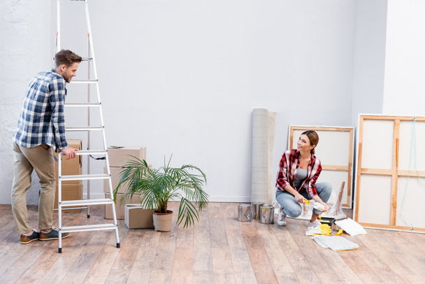 sourire jeune homme regardant femme avec rouleau de peinture tout en s'appuyant sur l'échelle à la maison - Photo, image