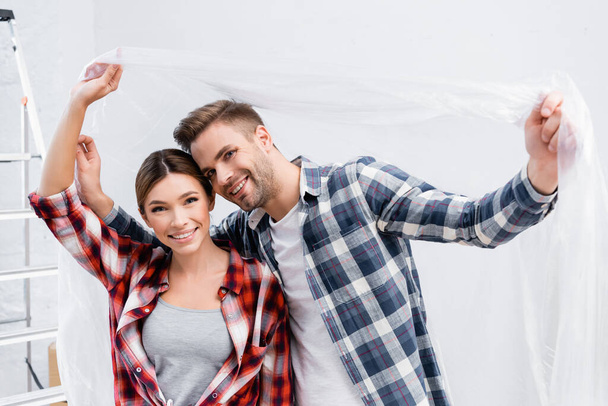 happy young couple looking at camera while holding polyethylene at home - Valokuva, kuva