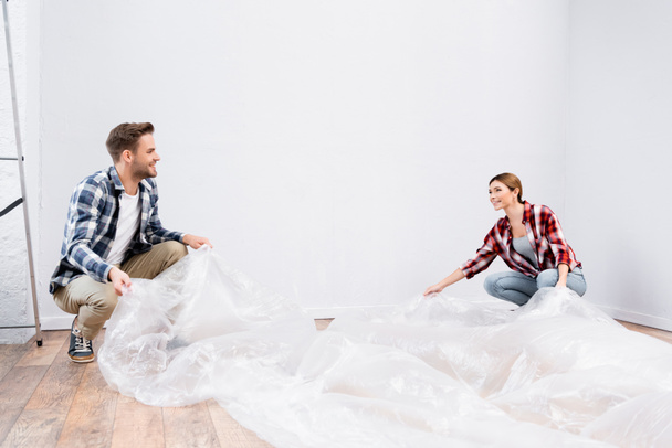 happy young couple with polyethylene covering room while sitting on floor at home - 写真・画像