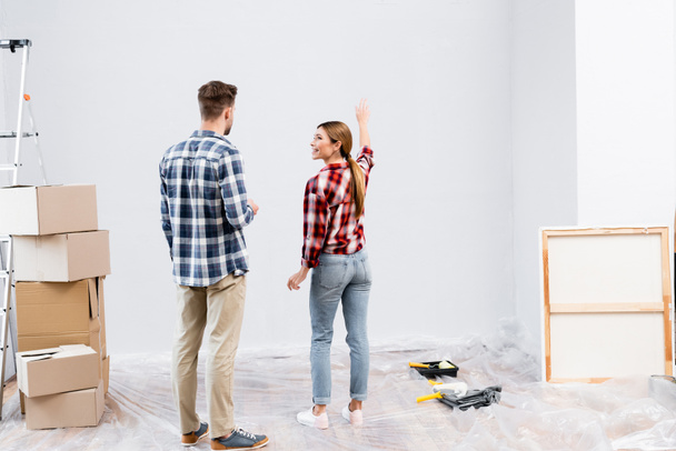 smiling young woman looking at man while pointing with hand at wall near picture, cardboard boxes and ladder at home - Foto, Imagem
