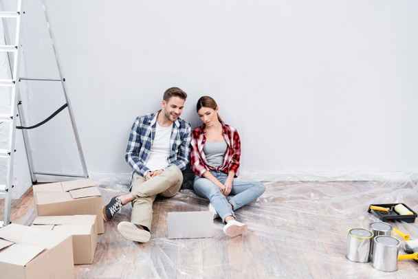 full length of happy young couple looking at laptop while sitting on floor near cardboard boxes, tins of paint and ladder at home - Foto, afbeelding