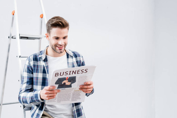 happy young man reading newspaper with business lettering near ladder indoors - Foto, afbeelding