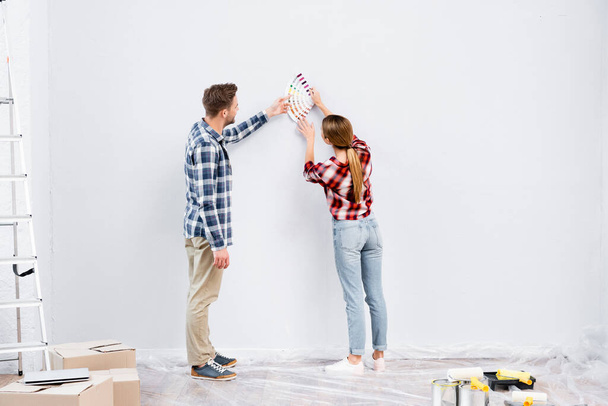 家での修理中に壁の色を選ぶパレットを持つ若いカップルの完全な長さ - 写真・画像