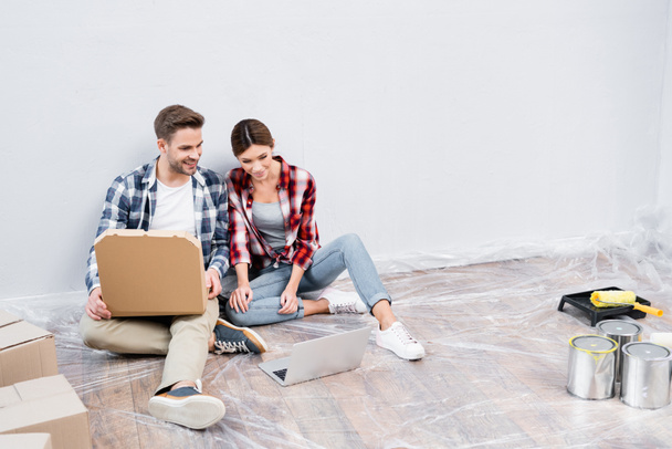 volledige lengte van glimlachende jonge man met kartonnen doos van pizza kijken film op laptop in de buurt van vrouw terwijl zitten op de vloer thuis - Foto, afbeelding