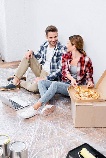 heureux jeune couple regardant l'autre tout en étant assis sur le sol près de la boîte de carton avec pizza à la maison - Photo, image