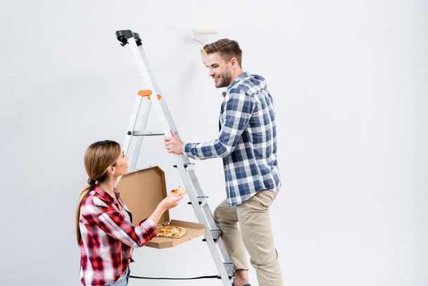 hymyilevä nuori mies rulla maalaus seinään katsomalla nainen antaa pala pizzaa lähellä tikkaat eristetty harmaa - Valokuva, kuva