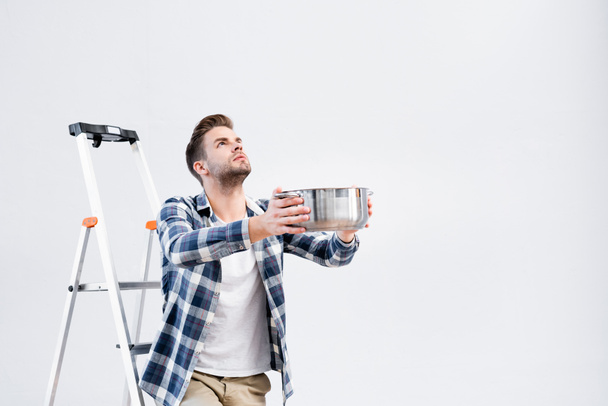 young man looking up while holding pot near ladder under leaking ceiling - Photo, Image