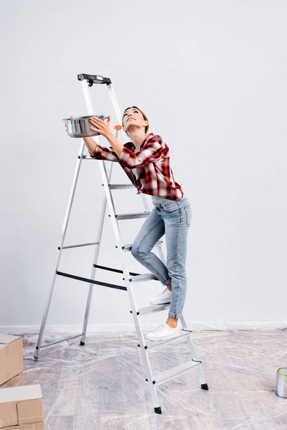 полный рост молодая женщина смотрит вверх, держа кастрюлю на лестнице под выщелачивающимся потолком дома - Фото, изображение