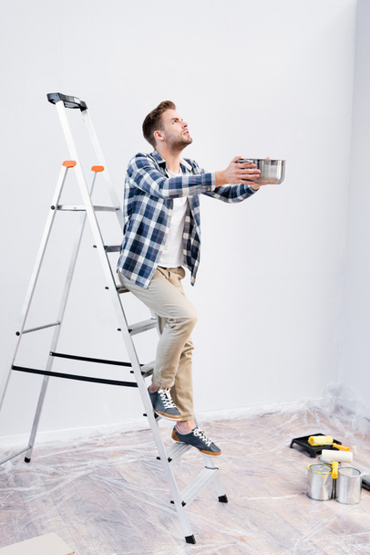 full length of young man looking up while holding pot and standing on ladder under leaking ceiling at home - Photo, Image