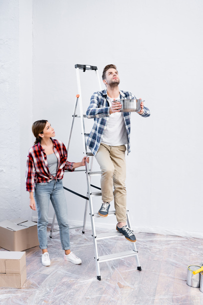 plná délka mladého muže drží pot, zatímco stojí na žebříku v blízkosti ženy pod tekoucí strop doma - Fotografie, Obrázek