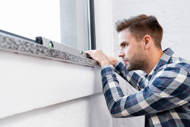 jeune homme concentré tenant outil de mesure de niveau sur le rebord de la fenêtre à la maison - Photo, image