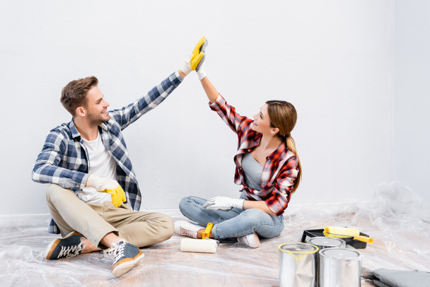 full length of happy young couple in gloves giving each other high five while sitting on floor near tins of paint at home - Valokuva, kuva