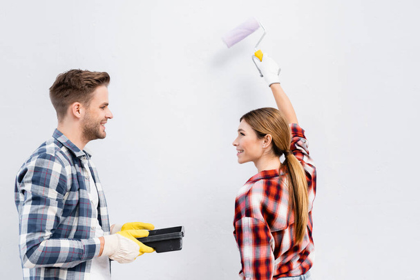 smiling young woman with paint roller looking at man holding tray isolated on grey - Foto, Imagem