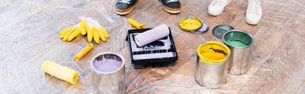 cropped view of couple feet near tins with paint, gloves, rollers and tray on floor covered with polyethylene, banner - Foto, Imagen