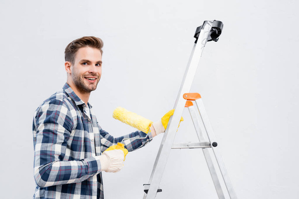 happy young man looking at camera while holding paint roller on ladder isolated on white - Valokuva, kuva