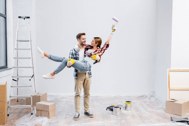 full length of happy young man lifting woman with paint roller at home - Foto, immagini